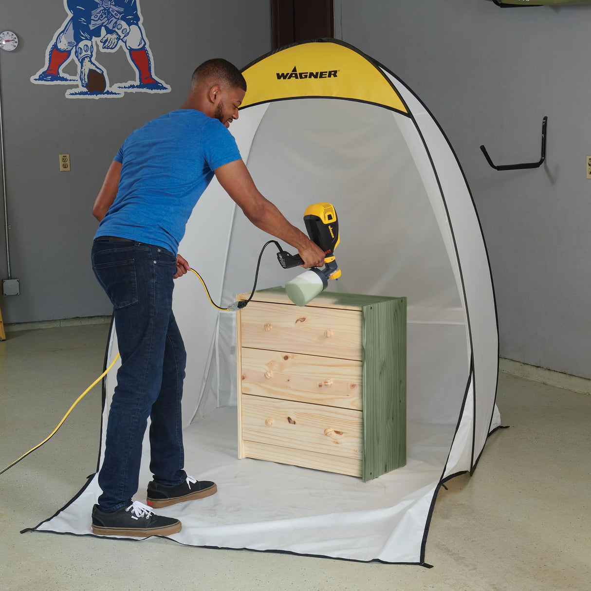 Person using a Wagner paint sprayer on a wooden box inside a portable spray booth.